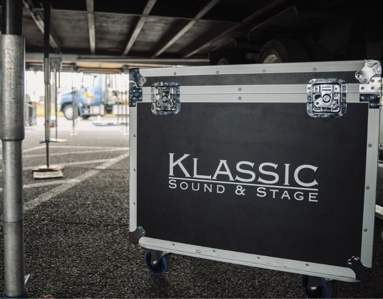 AV road case sitting under a stage while team is setting up for an outdoor concert.